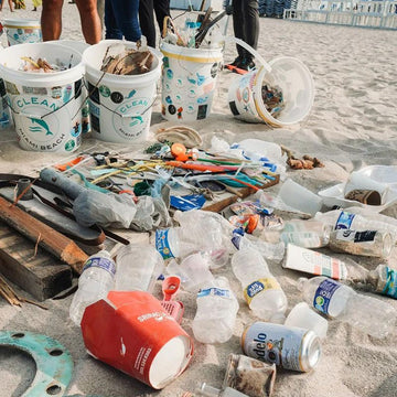 Beach littered with collected trash beside 'Clean Miami Beach' buckets.