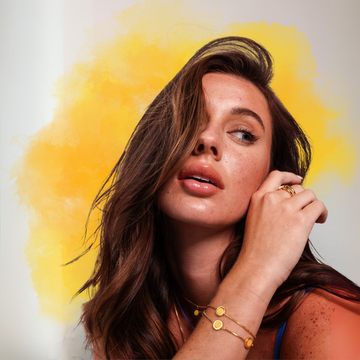 A woman with brown hair and freckles wearing Jason Hyde gold jewelry against a yellow background.