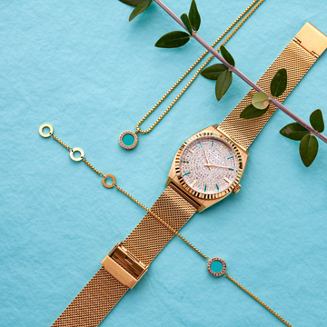 Gold-tone watch with mesh strap, matching necklace, and bracelet, on a blue background.
