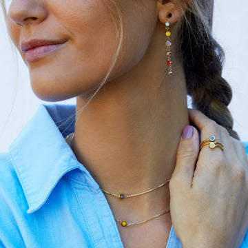 Layered gold necklaces and stacking rings with white and amber recycled ocean plastic details on blue shirt.