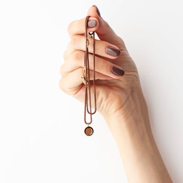 A hand with brown nail polish holds a Ocean Painted Brass Necklace
