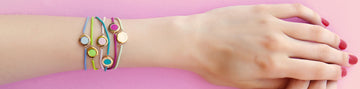 A hand wearing a stack of colorful beaded Ocean bracelets on a pink background.
