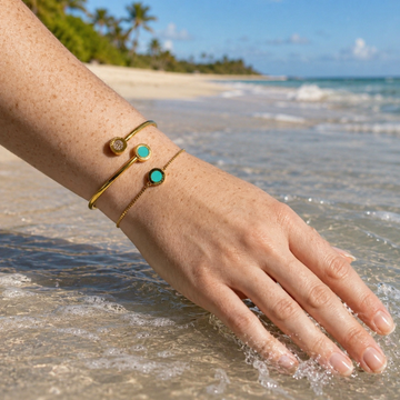 Two gold bracelets with turquoise and diamond-like discs on a persons arm dipped in ocean water