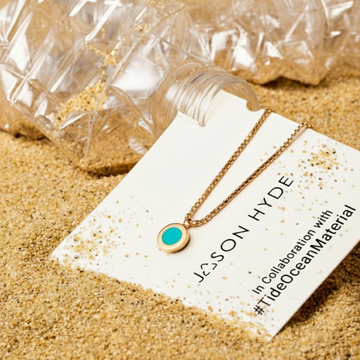 Gold-tone chain necklace with a small round pendant featuring a bright turquoise center on a white tag in sand