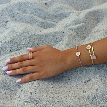 Gold ring and bracelets with pink enamel details on a persons hand on a sandy beach