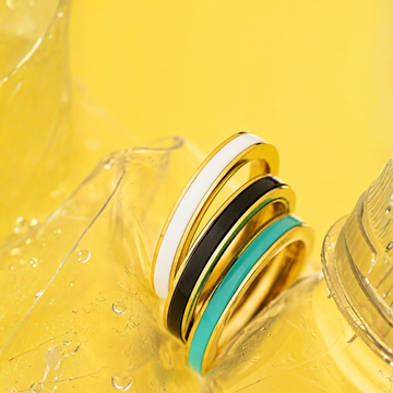Three gold stackable rings with white black and teal enamel bands styled on a yellow background