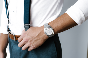 A man wearing a white shirt teal suspenders brown belt and a silver watch with a gray textured strap