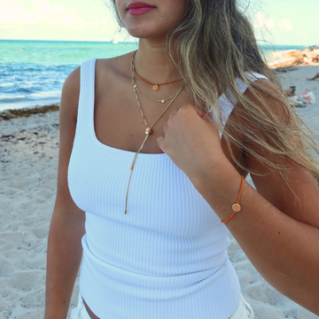Layered gold-tone necklaces with orange coin charms and an orange bracelet on a woman at the beach