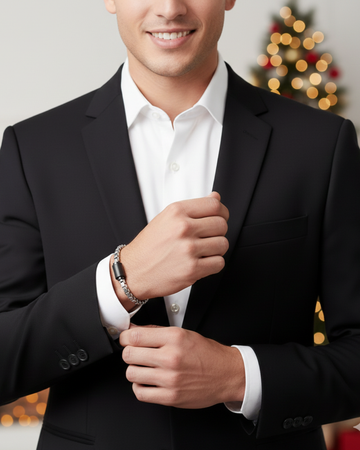 Close-up of a smiling man in a black suit and white shirt, adjusting his cuff while wearing a silver chain bracelet 