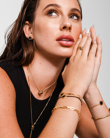 Woman wearing different black and gold jewelry pieces, wewa triple chain necklace, ocean bracelets, italy bracelet, new wave bracelet and bangle, black ocean earring. Woman is looking to the side