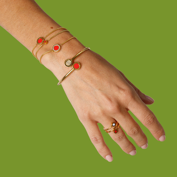 Gold jewelry with red enamel and zirconia on a woman's hand, against a green background.