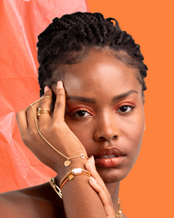 A woman with braided hair wearing a Jason Hyde gold-tone cuff bracelet with a beige charm and a delicate chain bracelet
