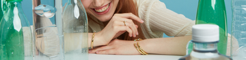 Close-up of a model smiling brightly, featuring stacked gold-tone bracelets and rings, set against a light blue background with water bottles.