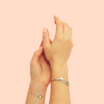 Two hands wearing delicate silver bracelets with small heart charms