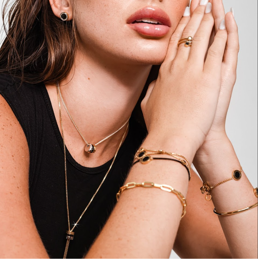 Woman wearing gold and black jewelry: earrings, necklaces, bracelets, ring.