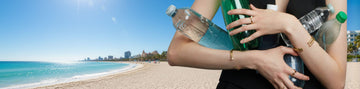 Hands holding plastic bottles on a sunny beach with buildings in the background.