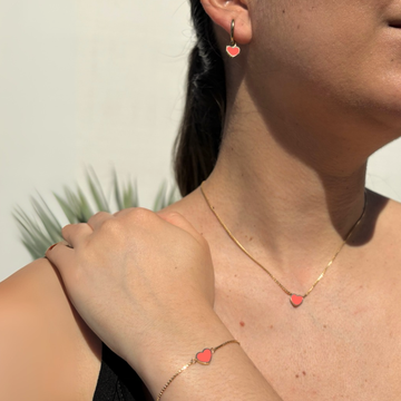 Model wearing a matching gold chain necklace, bracelet, and earring set featuring delicate coral-colored heart charms by Jason Hyde.