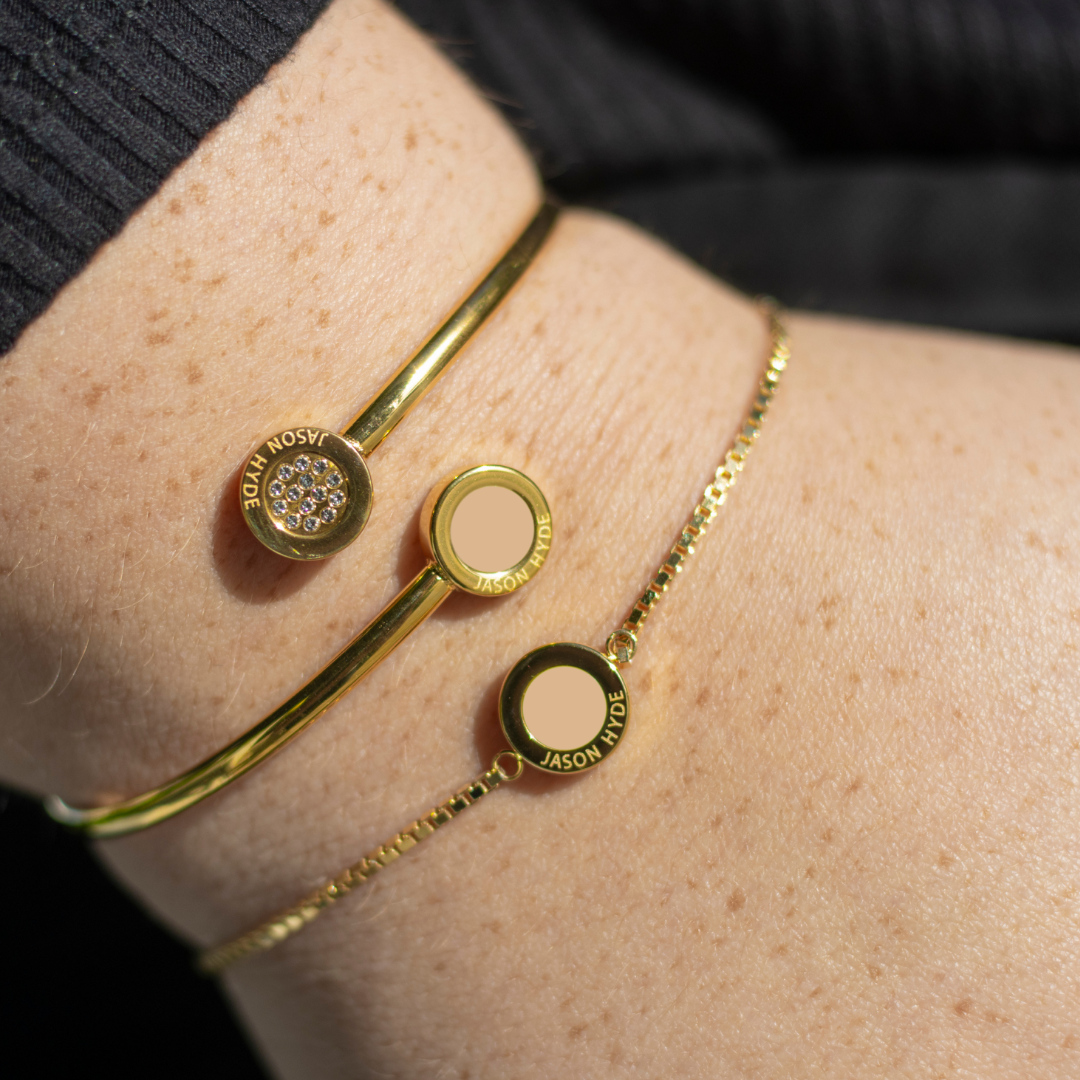 Wrist wearing a gold Jason Hyde pave bangle and a gold bracelet with a beige chip.