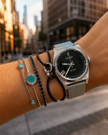 Person wearing a silver mesh watch with stacked black and turquoise stone bracelets outdoors on a city street.