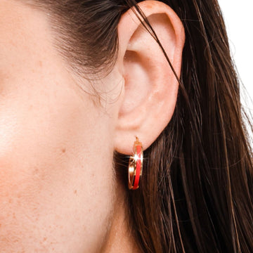 A close-up shot of a person's ear wearing a modern gold hoop earring with a vibrant red enamel stripe