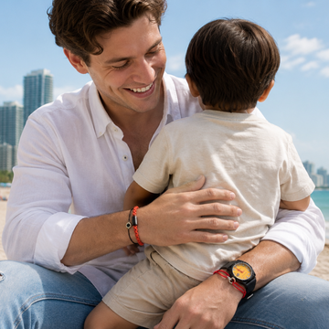 A dad in a white shirt and jeans sits on a beach, warmly hugging his young son