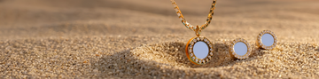 Jason Hyde gold round pendant necklace and matching stud earrings with crystal halo on beach sand.