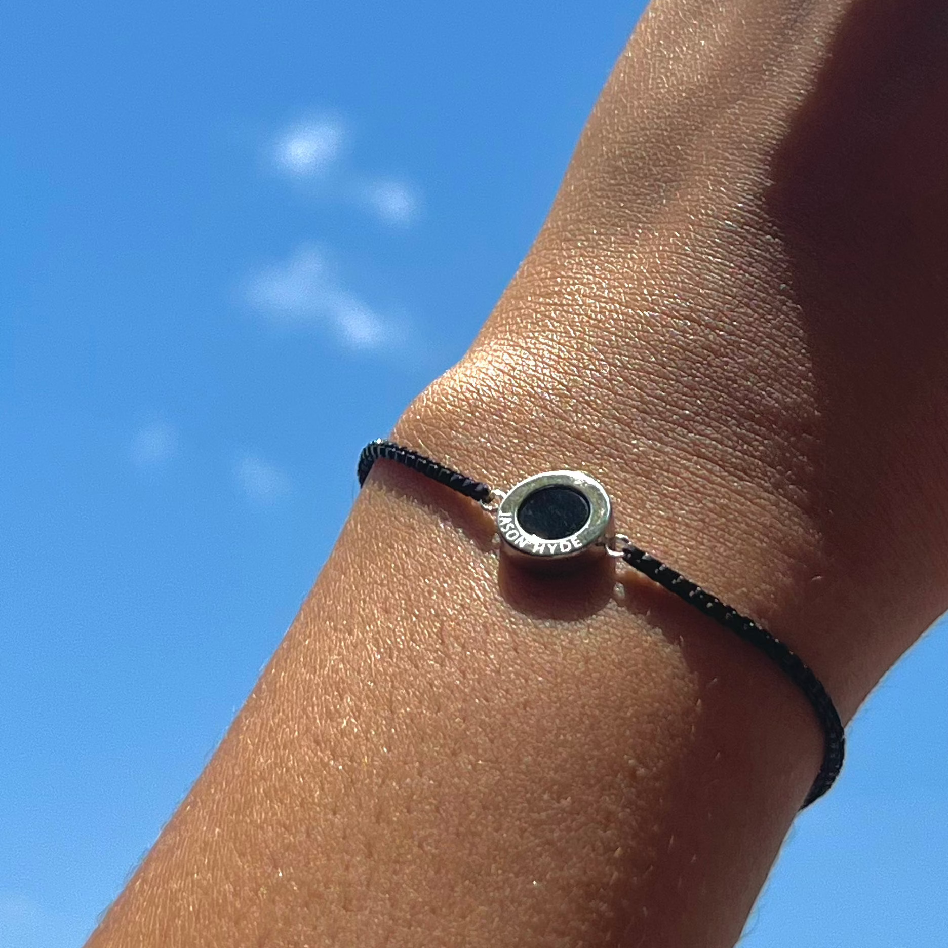 Wrist shot of Jason Hyde black cord bracelet and black pendant against blue sky.