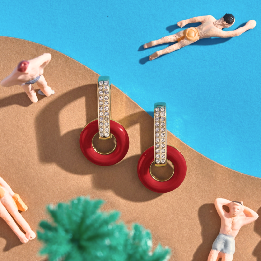 Red circular earrings with zirconia accents, displayed in a miniature beach