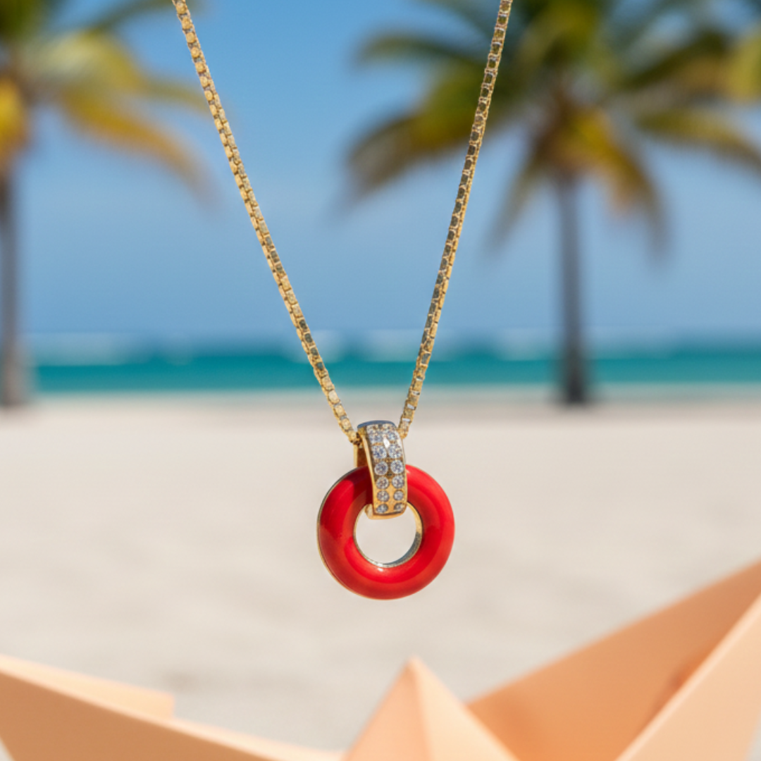 Gold necklace with a red circle pendant and zirconia accents on a beach background.