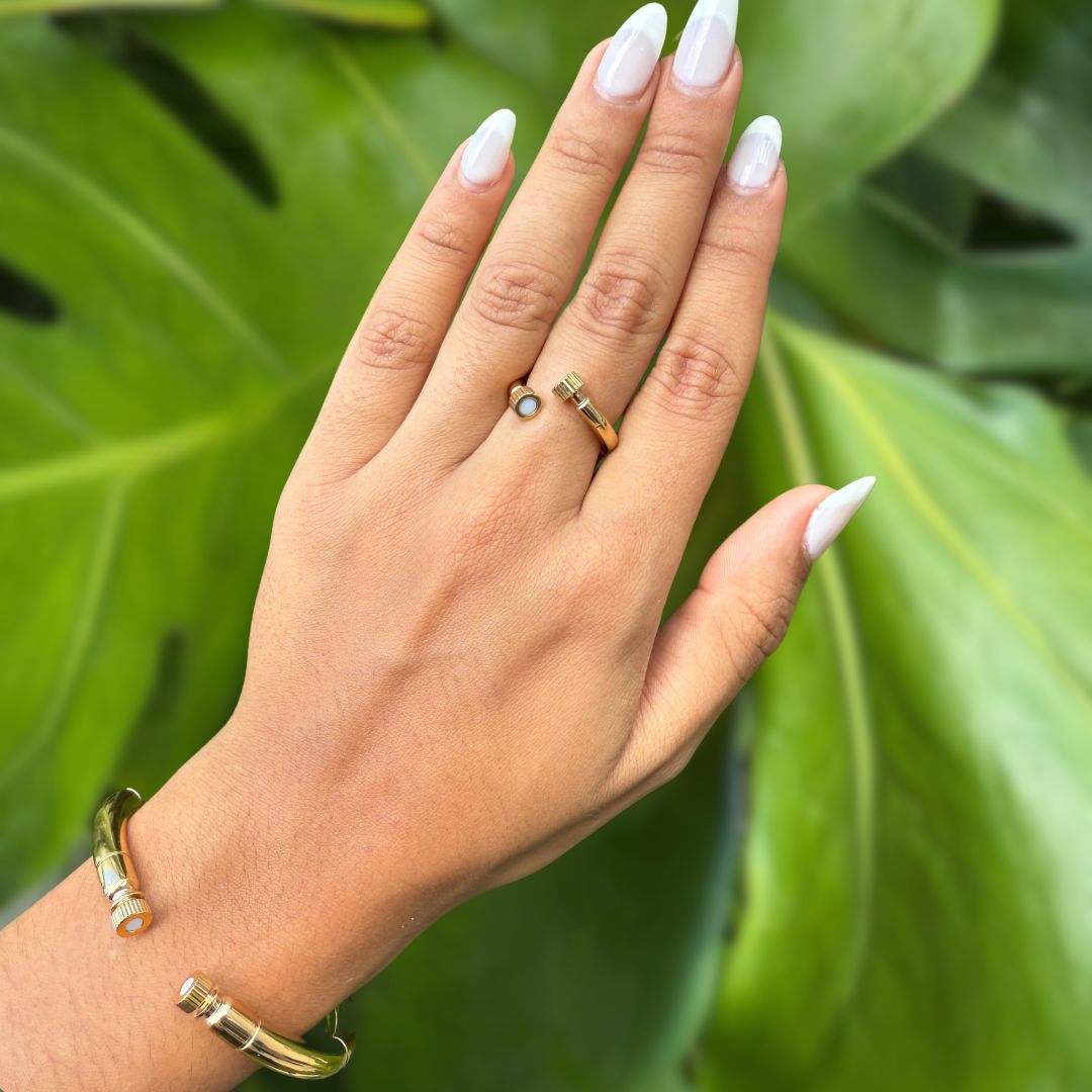 A hand wearing a gold open ring with a white chip and a gold cuff bracelet with cylindrical ends
