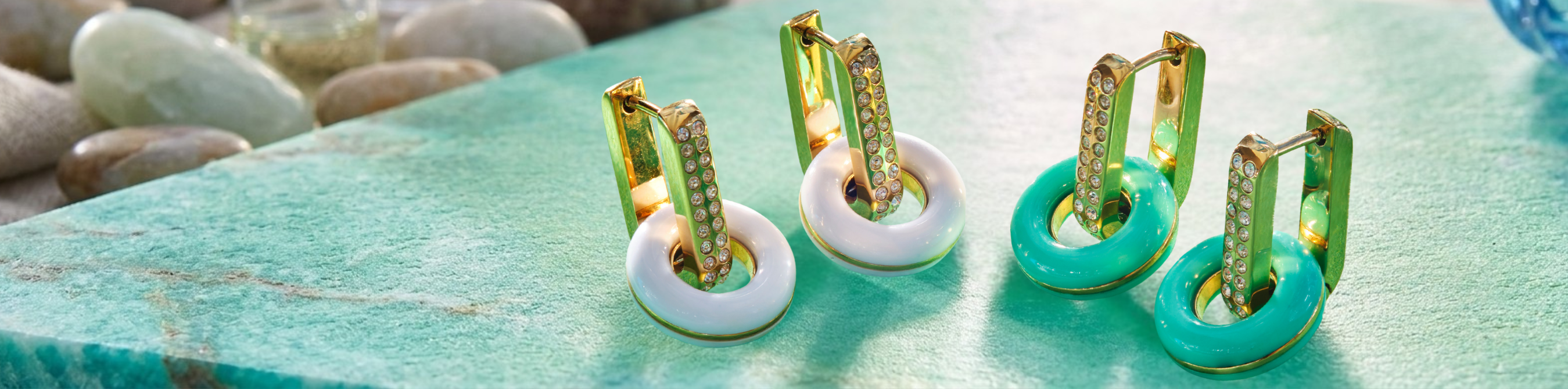 Jason Hyde luxury huggie earrings in gold with white and turquoise recycled plastic charms on a teal stone surface.