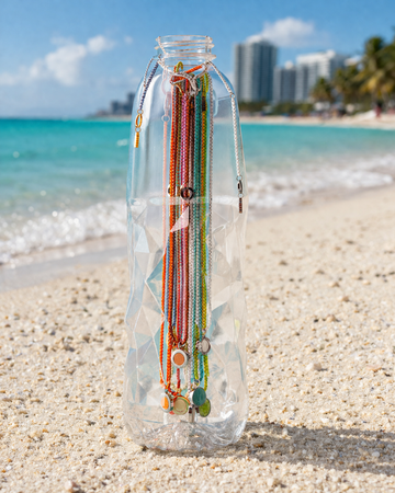 Colorful Jason Hyde beaded necklaces hanging inside a clear recycled plastic bottle on a sandy beach.
