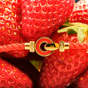 Red cord bracelet with a gold clasp and crystal details, laid over fresh strawberries.
