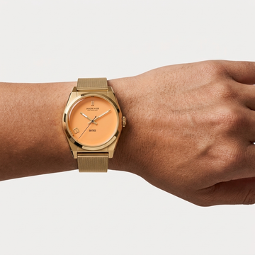 A person wearing a gold Jason Hyde watch with an orange dial and mesh strap.