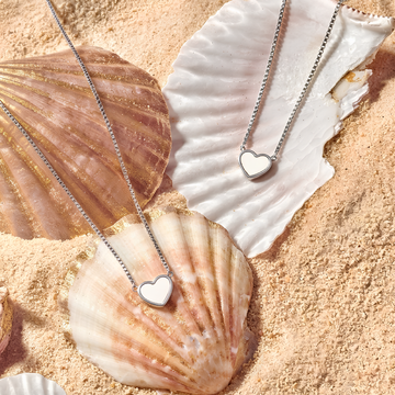 Two silver-colored stainless steel Jason Hyde heart necklaces resting on seashells in sand.