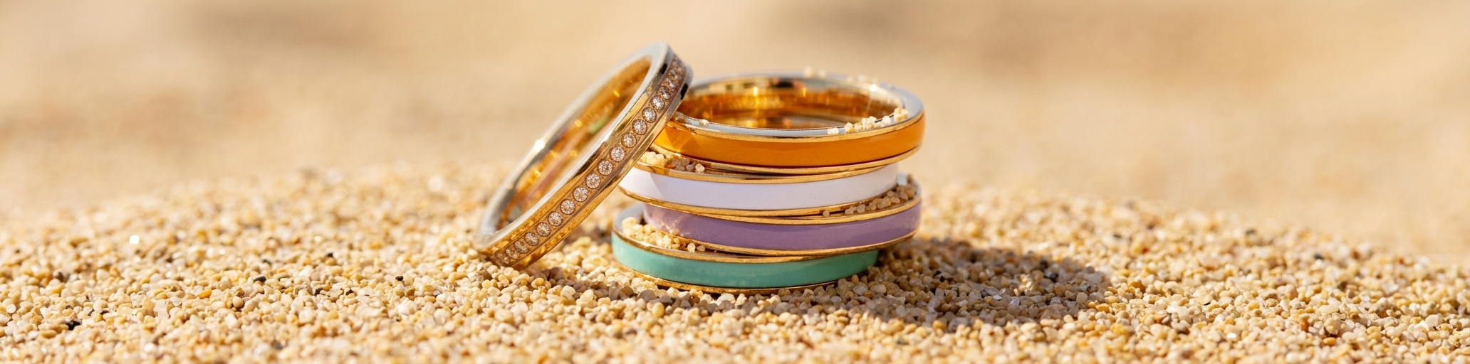 Stacked gold band rings with orange, white, purple, and turquoise enamel and one full-diamond pavé ring, resting on light sand.