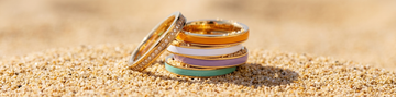 Stacked gold band rings with orange, white, purple, and turquoise enamel and one full-diamond pavé ring, resting on light sand.
