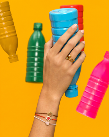 Woman's hand wearing a gold ring and colorful cord bracelets, holding a plastic bottle 