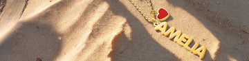 Custom gold name necklace 'AMELIA' with a red heart charm resting on beach sand with shadows.