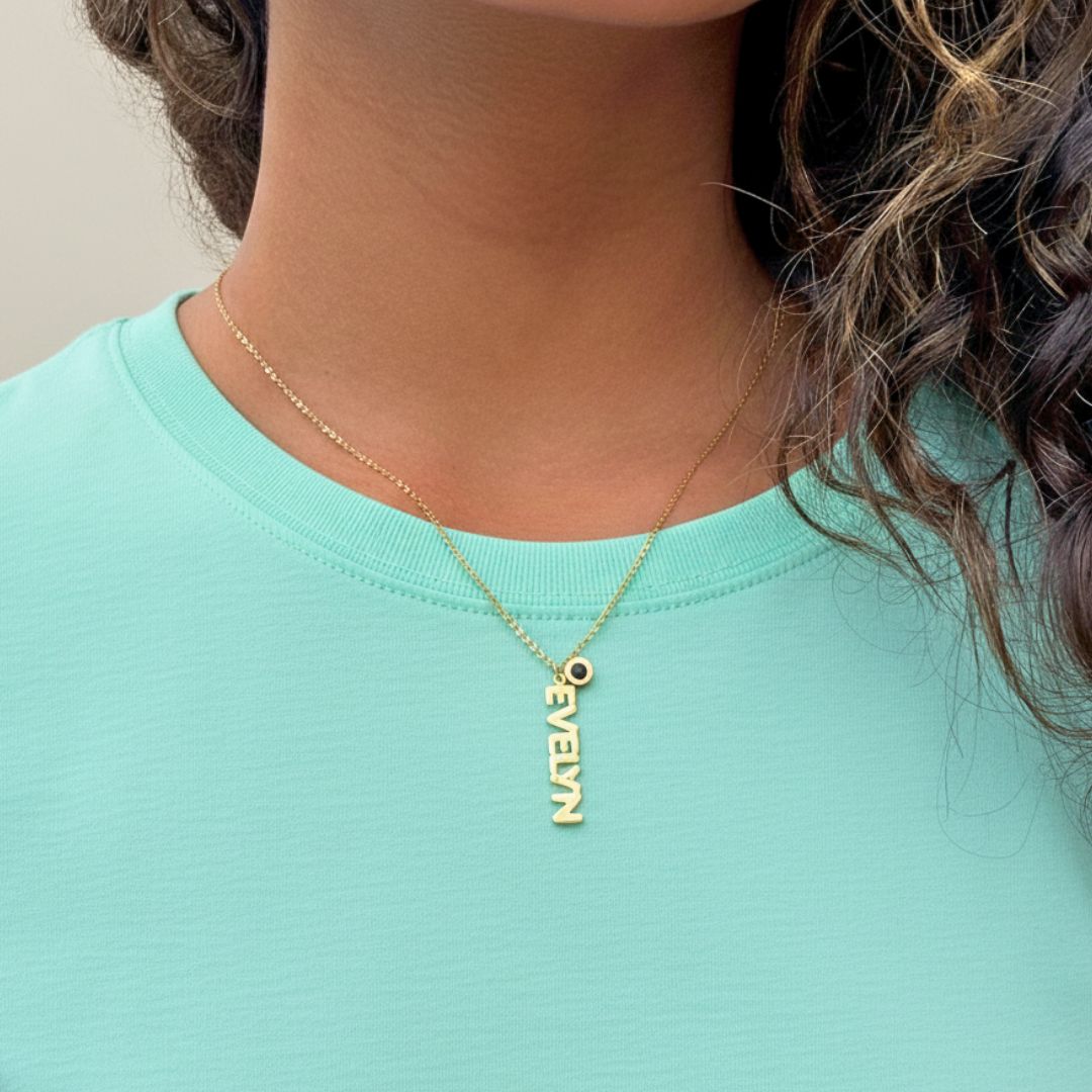 Gold vertical name necklace with "EVELYN" and a small black charm on a woman's neck.