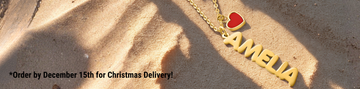 Custom gold name necklace 'AMELIA' with a red heart charm resting on beach sand with shadows.