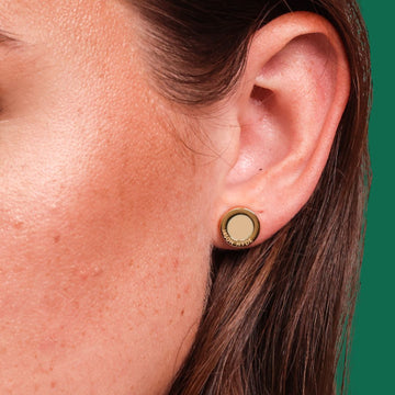 A close-up of a woman's ear wearing a round gold stud earring with a beige chip