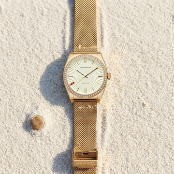 Elegant Jason Hyde gold mesh strap watch with a diamond-studded bezel, displayed on white beach sand.