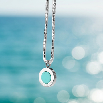 Jason Hyde silver necklace with a turquoise round pendant against a blurred ocean background.