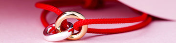 A close-up of a red cord bracelet with two interlocking ring charms, one gold and one silver.